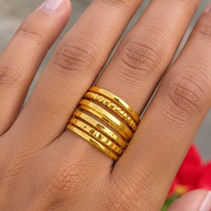 May include: A set of five stacked gold-toned rings worn on a finger. The collection includes one smooth band, one twisted rope-textured band, and three additional smooth bands of varying thicknesses, all featuring a polished metallic finish.