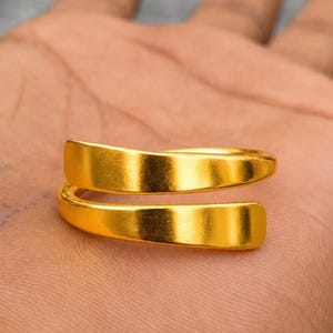 Puede incluir: Un anillo tipo bypass de color dorado con un acabado liso y pulido. La banda consta de dos extremos ligeramente curvados que se superponen alrededor del dedo, creando un diseño abierto y ajustable con una superficie metálica reflectante.