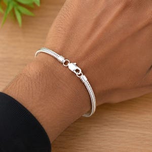 May include: A silver-toned wheat chain bracelet featuring a polished finish and a secure lobster claw clasp. The bracelet has a woven, textured link design that wraps around the wrist, showcasing a consistent metallic sheen throughout the piece.
