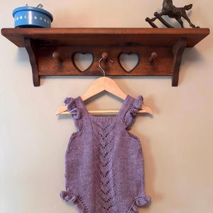 Könnte beinhalten: Ein lavendelfarbener Strickstrampler mit einem zentralen vertikalen Spitzenmuster, gerüschten Schulterträgern und gerüschten Beinöffnungen. Das Kleidungsstück hängt an einem hellen Holzkleiderbügel an einer Wandgarderobe.