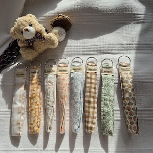 Peut inclure: Une collection de porte-clés en tissu avec divers motifs floraux et à motifs, chacun avec un anneau en métal. Les porte-clés sont étiquetés "Hand Made". Un petit ours en peluche et une balle de baseball avec un chapeau de gland sont également présents.