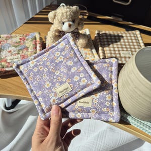 May include: Square, handmade coasters with floral patterns. The coasters feature a lavender background with small white and yellow flowers. Each coaster has a stitched border and a small "Hand Made" tag. A teddy bear and other coasters are in the background.