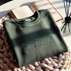 May include: An olive green sweatshirt with the embroidered text "support your local library." The sweatshirt is folded and resting on a light pink knitted cushion. An open book and a glass diffuser are in the background.