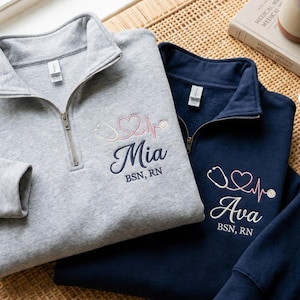 Może przedstawiać: Dwie bluzy z zamkiem błyskawicznym do połowy szyi, jedna szara i jedna granatowa, z haftem w postaci stetoskopu i linii bicia serca. Szara bluza ma napis "Mia", a granatowa "Ava", obie z napisem "BSN, RN" poniżej.