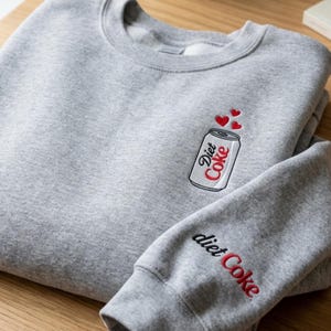 Op de afbeelding: Een grijs gemêleerd sweatshirt met ronde hals en een geborduurde afbeelding op de borst van een zilverkleurig frisdrankblikje met het label Diet Coke en drie kleine rode hartjes erboven, plus de tekst Diet Coke geborduurd op de mouw.