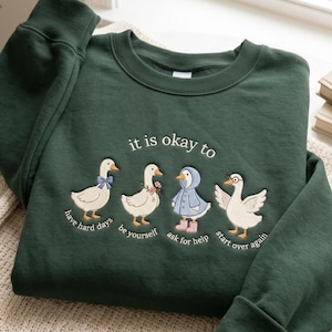 Puede incluir: Sudadera verde oscuro con patos bordados y la frase "it is okay to". Debajo de los patos están las frases "have hard days", "be yourself", "ask for help" y "start over again".