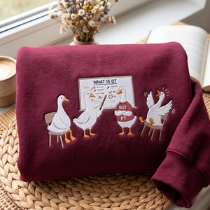 May include: A deep burgundy sweatshirt featuring an embroidered graphic of four white geese in a classroom setting. One goose points to a whiteboard labeled WHAT IS OT, while another wears a red shirt with the text NOT PT. The design includes chairs.
