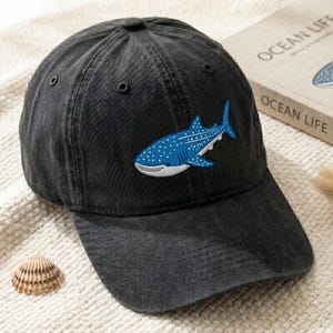 May include: A black baseball cap with a curved brim. The cap features a blue and white embroidered whale shark patch on the front. The text "OCEAN LIFE" is visible on a book in the background.