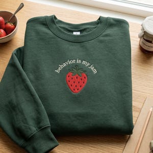 Pode incluir: Uma camisola de gola redonda em verde floresta escuro com um gráfico de morango vermelho bordado no centro do peito. Acima do morango, o texto branco "behavior is my jam" está escrito em minúsculas e em arco. A peça tem mangas compridas.