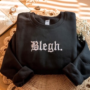 以下が含まれることがあります： 白いゴシック体で「Blegh.」と刺繍された黒いスウェットシャツ。スウェットシャツは、背景に本と時計がある、織られたマットの上に表示されています。この衣服はクルーネックと長袖です。
