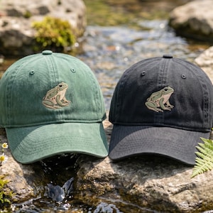 May include: Two baseball caps, one in forest green and one in black, each featuring an embroidered graphic of a green frog on the front panel. The caps have a structured crown, a curved brim, and a button at the top center of the fabric dome.