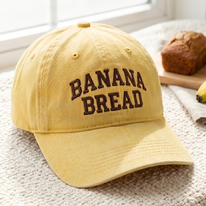 May include: Yellow baseball cap with the words "BANANA BREAD" embroidered in brown. The cap has a curved brim and a button on top. A loaf of banana bread is in the background.