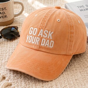 May include: A peach-colored baseball cap with the embroidered text "GO ASK YOUR DAD" in white. The cap has a curved brim and a small button on top. The hat is sitting on a textured surface.