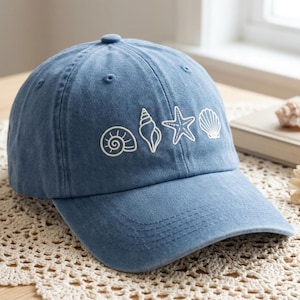 May include: A blue denim baseball cap with white embroidered seashell designs. The cap features a curved brim and a classic six-panel design, perfect for casual wear and beach outings.