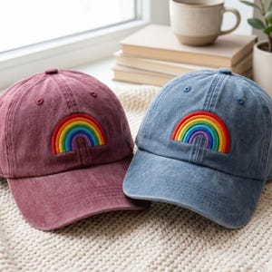 May include: Two baseball caps, one in a muted maroon and the other in a faded blue, each featuring a vibrant, embroidered rainbow patch on the front panel. Both caps have a structured crown, a curved brim, and matching stitching details.