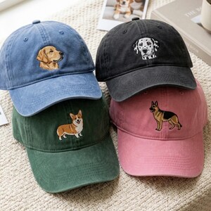 May include: A collection of four baseball caps in blue, black, green, and pink. Each cap features an embroidered dog design on the front panel: a golden retriever, a dalmatian, a corgi, and a German shepherd, respectively.
