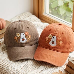 May include: Two baseball caps, one brown and one orange, each featuring an embroidered design of a white ghost holding a small orange pumpkin, surrounded by yellow and orange flowers. The caps have a structured crown and a curved brim.