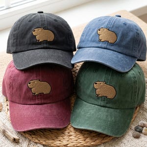 Op de afbeelding: Vier baseballpetten in antraciet, blauw, bordeauxrood en groen, elk voorzien van een geborduurde beige capibara op het voorpaneel. De petten hebben een verwassen textuur, een gebogen klep en een knoopje bovenop de kroon van de pet.