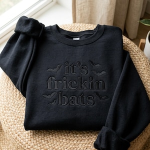 May include: A black long-sleeve crewneck sweatshirt featuring tonal black embroidery that reads "it's frickin bats" in a centered serif font, surrounded by small embroidered bat silhouettes scattered across the chest area.