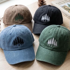 Può includere: Quattro cappellini da baseball marroni, neri, blu e verdi. Ogni cappellino presenta un ricamo di alberi. I cappellini sono realizzati in un tessuto morbido e strutturato e hanno una visiera curva. Sono esposti su una superficie di legno.