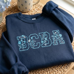Peut inclure: Sweat-shirt bleu marine avec les lettres "BCBA" brodées dans un motif floral bleu clair. Le sweat-shirt est plié sur une surface tissée, avec une tasse et un vase fleuris en arrière-plan.