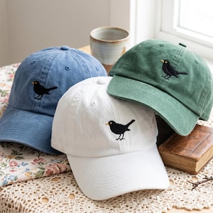 May include: Three baseball caps in blue, white, and green, each featuring an embroidered black bird with a yellow beak on the front panel. The caps have a structured crown, a curved brim, and stitched eyelets for ventilation on the top panels.