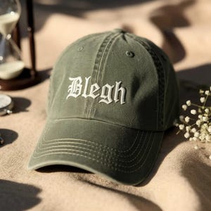 May include: Olive green baseball cap with the word "Bleigh" embroidered in white script across the front. The cap has a curved brim and a classic six-panel design. The hat is made of a soft, worn material.