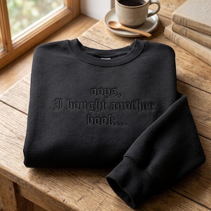 Puede incluir: Una sudadera negra de manga larga con el texto bordado "oops, I bought another book..." en una fuente de estilo gótico oscuro centrada en el pecho. La prenda cuenta con cuello redondo y puños acanalados en un diseño sencillo y limpio.