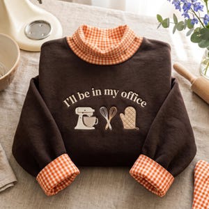 May include: A dark brown long-sleeve sweatshirt featuring an orange and white gingham mock neck collar and matching sleeve cuffs. The front displays the embroidered text "I'll be in my office" above icons of a stand mixer, whisks, and an oven mitt.
