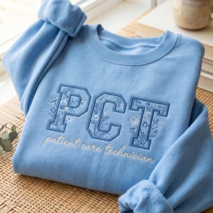 May include: A light blue sweatshirt with the embroidered letters "PCT" in a floral design. Below the letters, the words "patient care technician" are embroidered in a cursive font. The sweatshirt is folded on a woven surface.