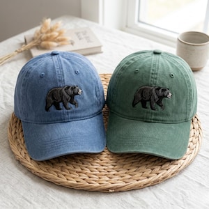 Puede incluir: Dos gorras de béisbol, una en azul desteñido y otra en verde apagado, cada una con un gráfico de oso negro bordado en el panel frontal. Ambas gorras tienen una corona estructurada, una visera curva y ojales cosidos para ventilación.