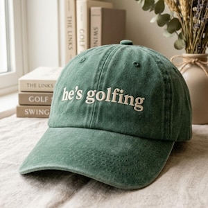 May include: A green baseball cap with the embroidered text "he's golfing" in white. The cap has a curved brim and a button on top. The hat is made of a soft, textured fabric.