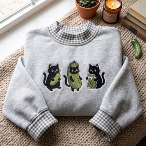Puede incluir: Una sudadera gris claro con cuello alto y puños con estampado de cuadros vichy en gris y blanco. La parte delantera muestra un bordado de tres gatos negros, uno sosteniendo un pepinillo, otro disfrazado de pepinillo y otro sosteniendo un frasco de pepinillos.