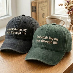 May include: Two baseball caps, one black and one green, with the phrase "inshallah-ing my way through life" embroidered in white. The caps are made of a worn, textured fabric, suggesting a vintage style. The caps are sitting on a wooden surface.