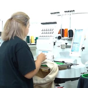 May include: A white industrial embroidery machine with multiple thread spools in various colors, including orange, black, yellow, and pink. The machine features a digital screen and is actively stitching a beige garment held in a circular green hoop.