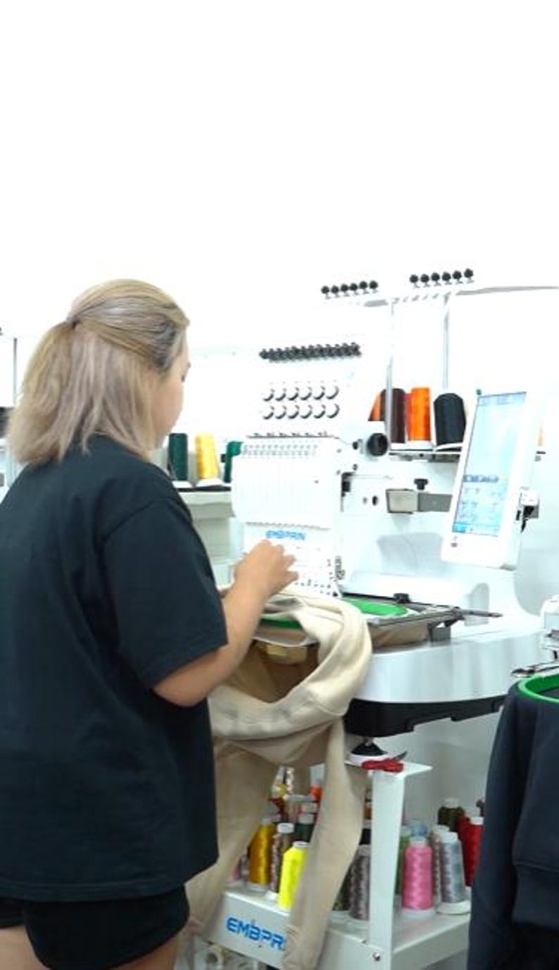 May include: A white industrial embroidery machine with multiple thread spools in various colors, including orange, black, yellow, and pink. The machine features a digital screen and is actively stitching a beige garment held in a circular green hoop.