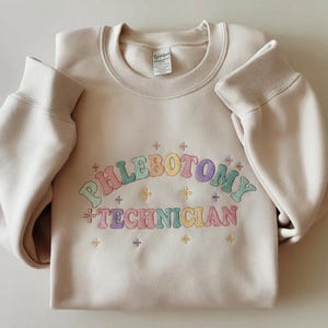 May include: A beige sweatshirt with the words "PHLEBOTOMY TECHNICIAN" embroidered in a rainbow of pastel colors. The sweatshirt has a crew neck and long sleeves with ribbed cuffs. Small cross-shaped designs are embroidered around the text.