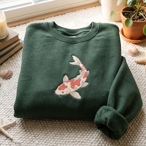 Peut inclure: Un sweat-shirt à col rond vert forêt foncé orné d'une broderie centrale représentant une carpe koï blanche avec des taches rouge orangé. Le vêtement possède des manches longues avec des poignets côtelés et un col côtelé en tissu uni.