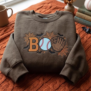 May include: A brown crewneck sweatshirt featuring an embroidered design on the chest. The design includes the orange letter B, a white baseball with red stitching, and a brown baseball glove, surrounded by orange spiderwebs and black bats.