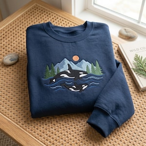 May include: A navy blue crewneck sweatshirt featuring an embroidered design of two orcas swimming in blue water. The scene includes green pine trees, light blue mountain peaks, and a small orange sun centered above the whales on the chest area.