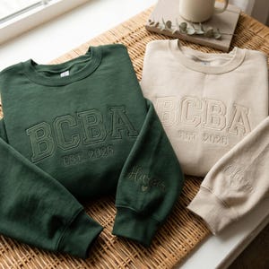 May include: A dark green sweatshirt and a beige sweatshirt, both featuring the embroidered text BCBA and EST. 2026 on the chest. The sleeves are personalised with the embroidered names Harper and Olivia, each accompanied by a small heart icon.