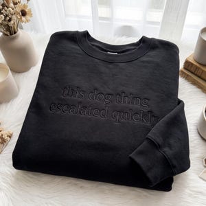 May include: A black long-sleeve sweatshirt featuring a crew neckline and ribbed cuffs. The front of the garment displays the embroidered text "this dog thing escalated quickly" in a matching black thread, creating a subtle, tonal appearance.
