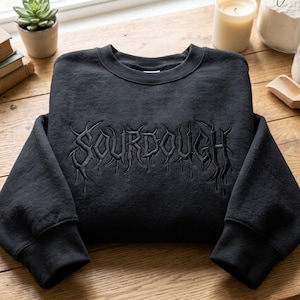 May include: A black crewneck sweatshirt featuring the word SOURDOUGH embroidered in a dark, stylized, thorny font across the chest. The garment has long sleeves with ribbed cuffs and a ribbed neckline, showcasing a monochromatic design.