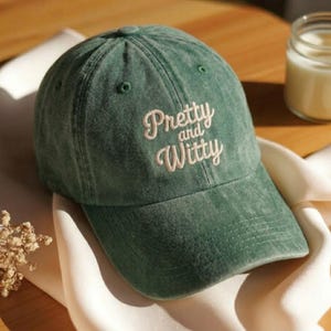 Puede incluir: Gorra de béisbol verde con el texto bordado "Pretty and Witty" en fuente cursiva. La gorra tiene una visera curva y un diseño clásico de seis paneles. La tela tiene un aspecto ligeramente descolorido.