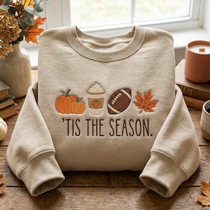 May include: A beige crewneck sweatshirt featuring embroidered autumn-themed icons including two orange pumpkins, a coffee cup with whipped cream, a brown football, and an orange maple leaf, with the text 'TIS THE SEASON. embroidered below.