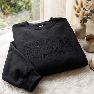 Könnte beinhalten: Ein schwarzes Sweatshirt mit Rundhalsausschnitt, das das Wort SOCIAL in großen, blockartigen Stickbuchstaben mit floralen Details im Inneren zeigt, sowie das Wort worker in Schreibschrift darunter, beides in passendem Schwarz.