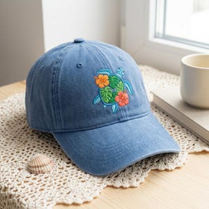 May include: A blue baseball cap featuring an embroidered design of a sea turtle with a shell composed of green monstera leaves, accented by two orange hibiscus flowers. The cap has a structured crown, a curved brim, and a button on top.