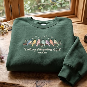 May include: A dark green crewneck sweatshirt featuring an embroidered design of seven colourful birds perched on a branch with musical notes above them. The text reads, "I will sing of the goodness of God" and "PSALM 89:1" in white lettering.