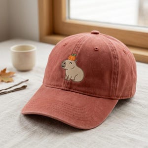 Puede incluir: Una gorra de béisbol color terracota con un bordado de un capibara marrón claro que lleva una pequeña calabaza naranja sobre la cabeza. La gorra cuenta con una visera curva, una corona estructurada con ojales cosidos y un botón superior.