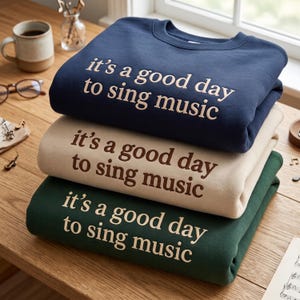 May include: A stack of three crewneck sweatshirts in navy blue, beige, and dark green. Each sweatshirt features the text "it's a good day to sing music" printed in a contrasting color on the front chest area, with long sleeves and a ribbed collar.